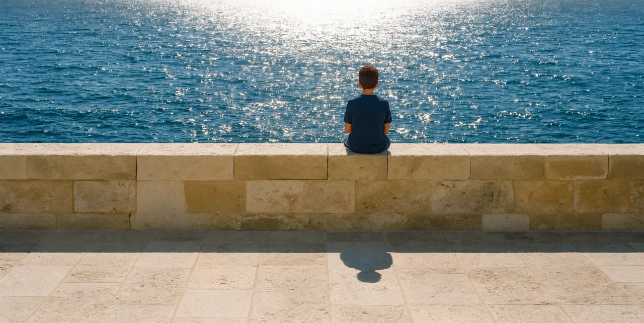 A boy sitting on a stone sea wall in Ortigia, Syracuse, looking out at the sunlit sea.