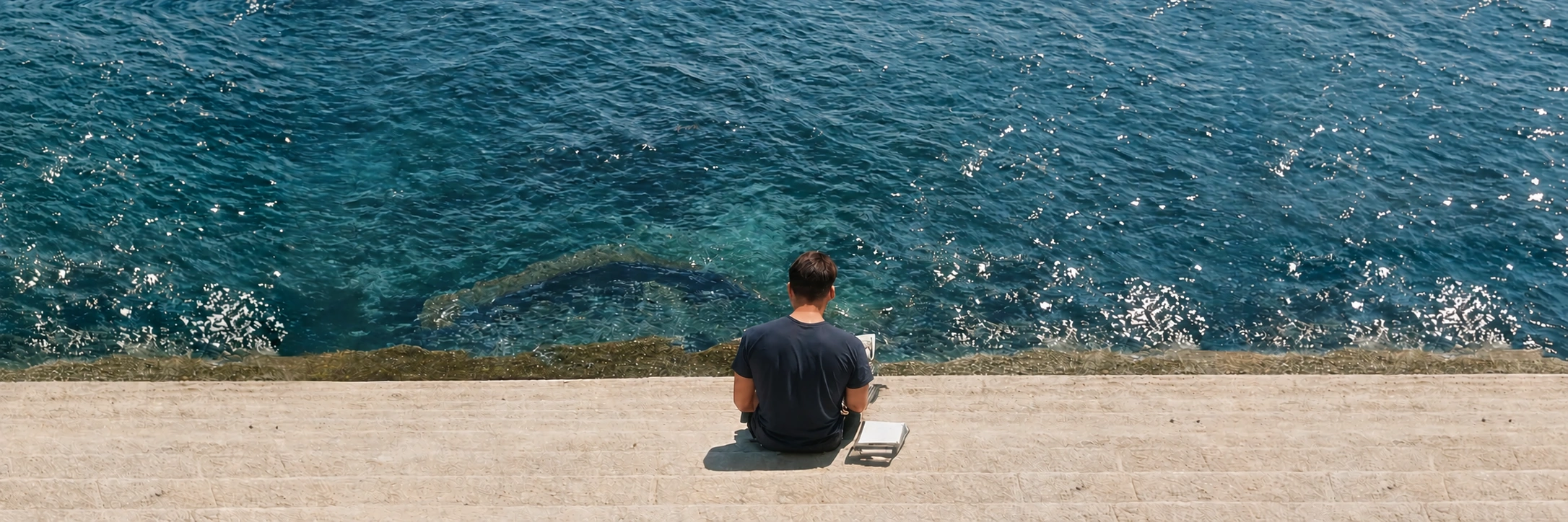 A young man seen from behind, sitting on stone steps by the sea in Ortigia, a book beside him.